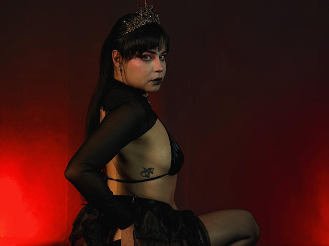 MadameDesadex with black mesh top, small black bikini bottom, tattoo on right side, tiara, red light, dark makeup, looking over shoulder, black skirt, sitting, showing her ass.