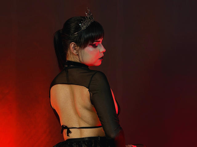 MadameDesadex with dark hair in a ponytail, wearing a sheer black top exposing her back, small ass, and a crown, standing in red and black lighting.