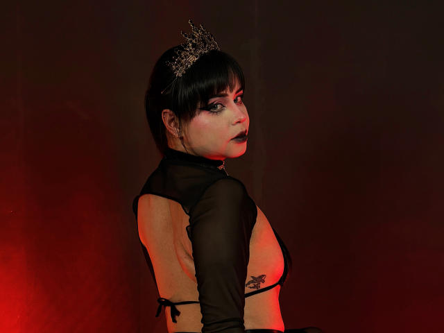 MadameDesadex with black lipstick, crown, and sheer black top, exposing her bare back and small tits, looking over her shoulder, red lighting on her pale skin.
