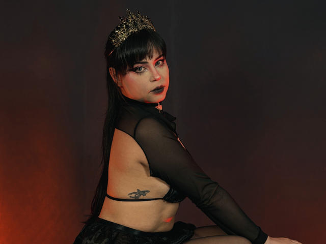 MadameDesadex with dark makeup, black sheer top, gold crown, and black skirt, showing her back tattoo and small black bra. Her ass is exposed, looking over her shoulder.