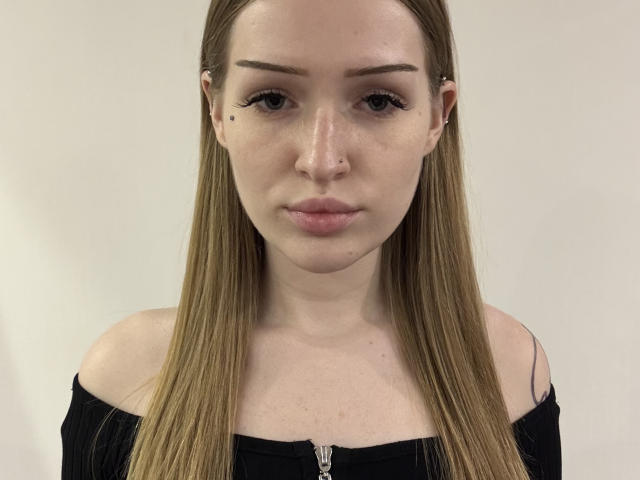 AbbieHalstead has long blonde hair, an off-shoulder black top, and a small mole on her right cheek. She has a nose ring and slightly pouted lips.