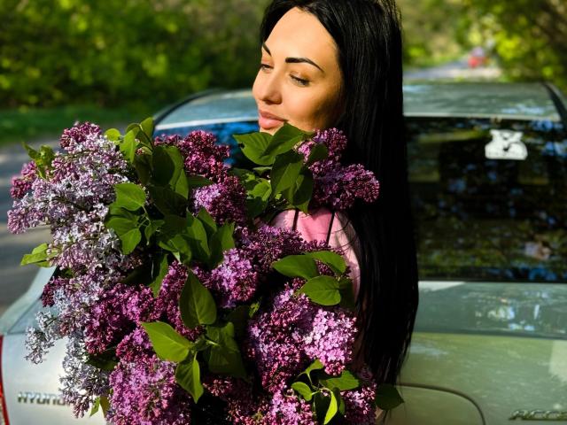 Xioni with long black hair, holding a large bouquet of purple and lilac flowers, standing in front of a silver car, looking down with a subtle smile.