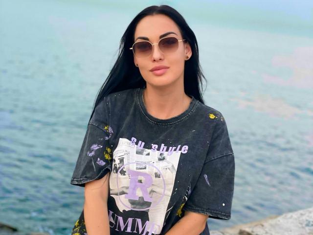 Xioni with long black hair, wearing a dirty grey t-shirt, round sunglasses, and pink lipstick, standing by a blue water body. Boobs are not visible.