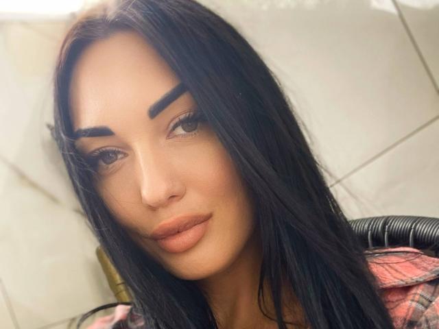 Xioni has dark, straight hair, thick eyebrows, and full, nude-colored lips. She's wearing a pink plaid shirt, looking at the camera with a slight, confident smile.