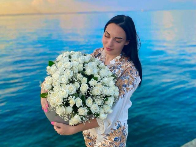 Xioni with white roses, holding a large bouquet, wearing a floral shirt, standing by blue water, long black hair, pink nails, looking down, arms around the flowers.