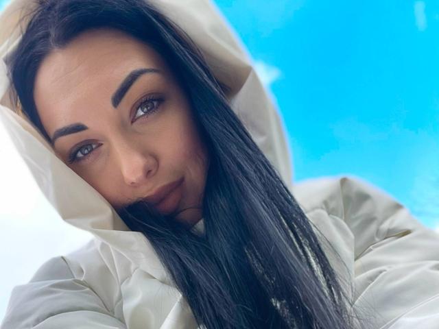 Xioni with long black hair, brown eyes, and dark eyebrows, wearing a white hoodie, looking slightly to the side with a subtle smile. Blue background.