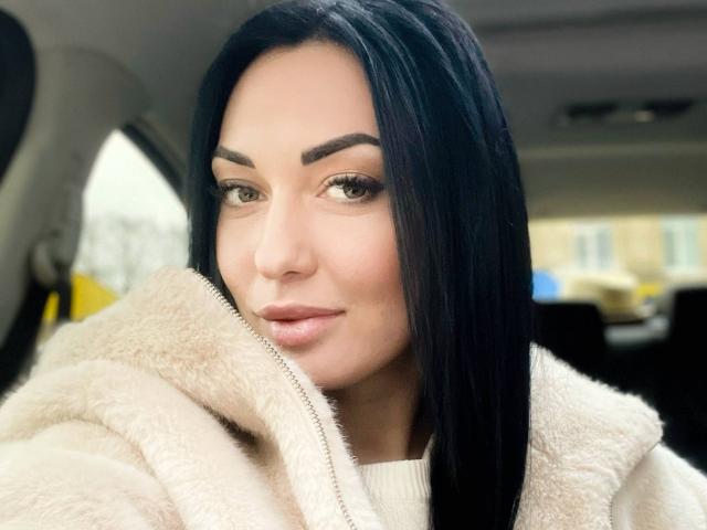 Xioni with long black hair, wearing a fluffy beige coat, has full eyebrows, subtle makeup, and a slight smile. Her tits are covered, and her pussy is not visible.