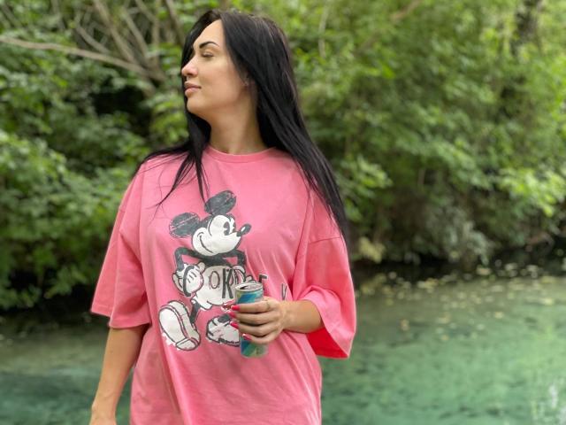 Xioni with long black hair, pink shirt with Mickey Mouse graphic, holding green bottle, standing by greenish water, looking to the side, casual, relaxed.
