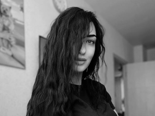 Black and white photo of Xioni with long, wavy dark hair covering one eye, wearing a black shirt, looking slightly to the side with a subtle, confident smile.