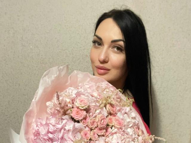 Xioni with long black hair, holding a bouquet of pink roses, has a subtle smile and is looking at the camera with slightly raised eyebrows.