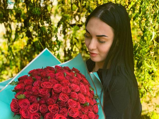 Xioni with long black hair holding a large bouquet of red roses, her eyes down, wearing a black shirt, standing in a sunlit green garden.