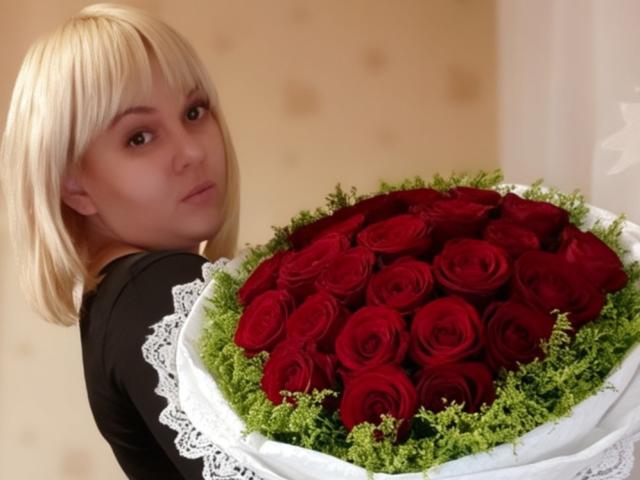 Leylisse with blonde hair holding a bouquet of red roses, her ass slightly turned, wearing a black top, white lace wrapping around the bouquet.