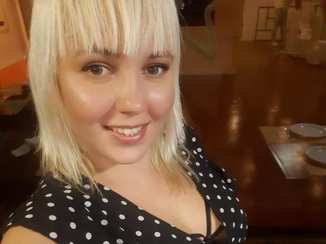 Leylisse with blonde hair, bangs, and a polka dot shirt, smiling at the camera, showing her shiny, cute face, and small tits, sitting at a wooden table.