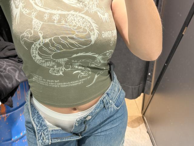 MargoFun's green tank top showing a snake graphic, blue jeans with white underwear, arm partially in frame, standing in a changing room.