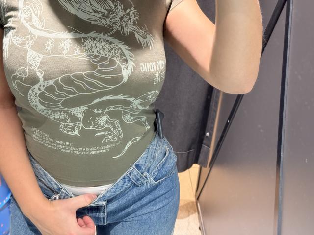 MargoFun's green t-shirt with white dragon design, showing her tits, tucked into blue jeans, with her hand on her white panties, taking a mirror selfie.