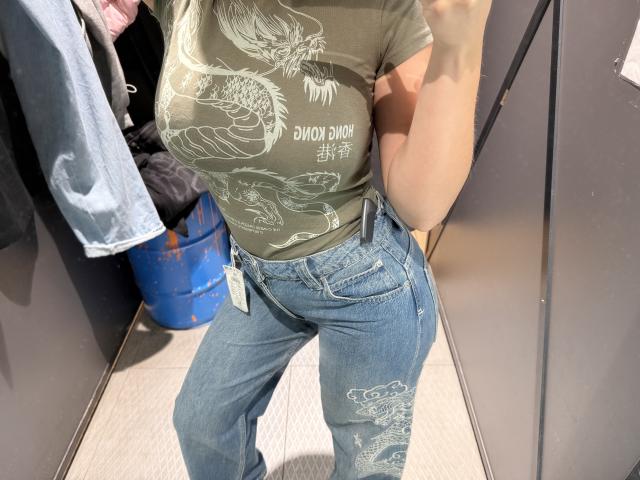 MargoFun with big boobs in a green graphic tee, blue ripped jeans, and a tag in the front pocket, standing in a fitting room, taking a selfie.