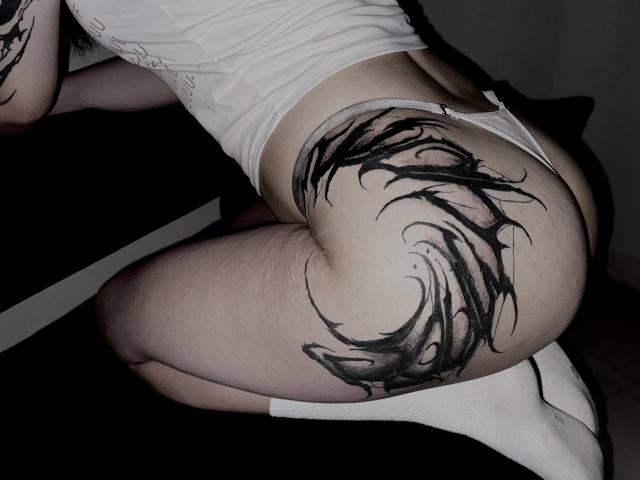 AnalAvi's white thong-ass showing a large black tribal tattoo on their pale, slightly hairy butt. They're kneeling with white socks, dark background, and a shadowy room.