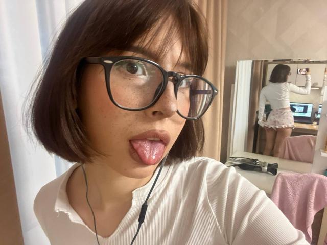 AnalAvi with short brown hair, black glasses, and tongue out, wearing a white ribbed shirt, reflected in a mirror, showing her ass in pink and white shorts.