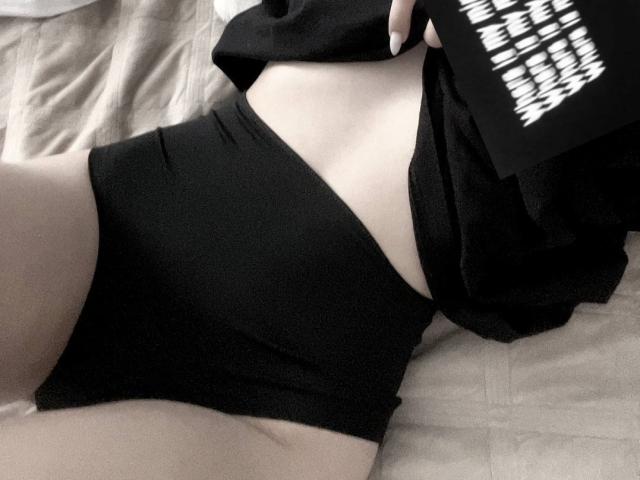 AnalAvi's black panties are snug on their pale, flat-ass, while they hold a black shirt over their midsection. A hand with white-painted nails holds the shirt.