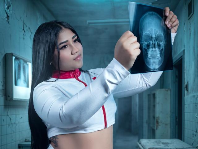 CamilaBosch met lang zwart haar, een witte crop top met een rode rits, die een röntgenfoto van een schedel vasthoudt, in een vervallen gang, die naar de camera kijkt, zelfverzekerd, aantrekkelijk, met zichtbare tatoeages aan de zijkant..