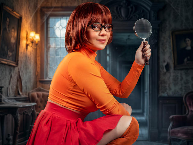 SherylHill with red bob, glasses, orange top, red skirt, holding magnifying glass, squatting, showing thigh, sexy, cute, indoors, vintage room, soft light, confident smile, nerdy, attractive.