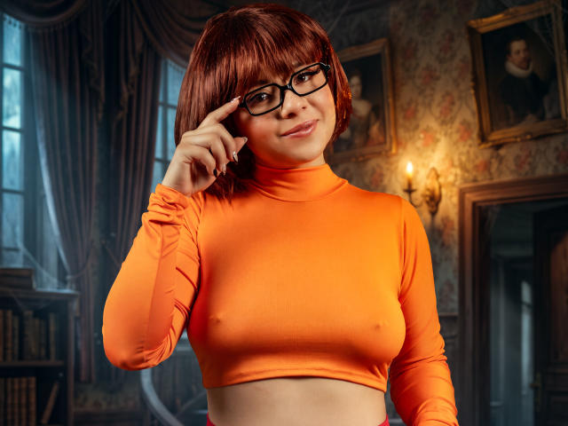 SherylHill with red bob, glasses, and orange crop top, showing her small tits, pointing at her glasses, smiling, in a vintage room with floral wallpaper.