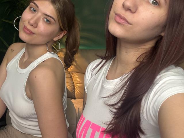 HotCaramelsTrio with brown hair, wearing a white tank top, showing tits, small hoop earrings, and pink text, in two mirrored selfies, looking cute, with a slightly pouty expression.