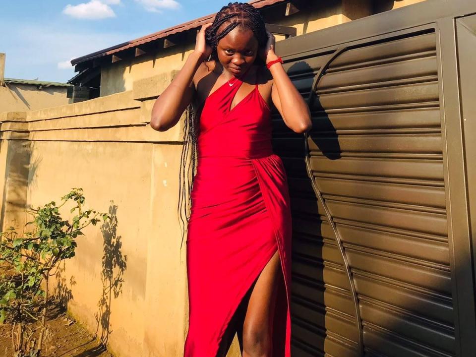 SavageBabe vestindo um vestido vermelho, sem mangas e com uma fenda alta, com as mãos nos cabelos trançados, em frente a um portão de metal, a luz do sol realçando sua pele escura e lisa..