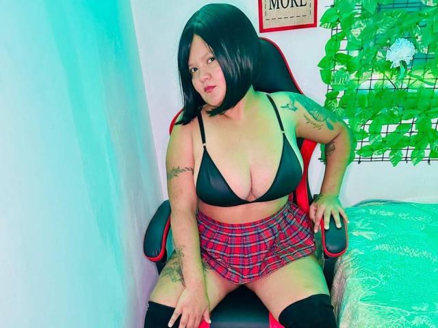 CamilaBacyc with black bob, wearing black bra, red plaid skirt, black thigh-highs, sitting on red-black chair, tattoos on arms, looking at camera, slightly smiling. Boobs visible.