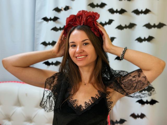 KaraCriegs mit braunen Haaren, lächelnd, trägt ein schwarzes Spitzenkleid, eine rote Blumenhaarreif, einen schwarzen Spitzenvelo und einen silbernen Armreif, Hände am Kopf, Brüste sichtbar, süß, sexy..