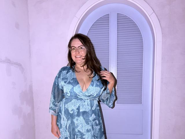 BadAngel with glasses, blue floral dress, medium tits, smiling, brown hair, holding hair, standing in front of arched shutter door, white walls, casual, confident.