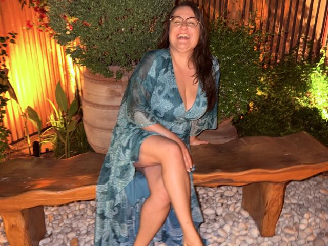 BadAngel with tits in a blue patterned robe, sitting on a wooden bench, legs crossed, wearing glasses, smiling slightly, in a greenery-filled, warmly-lit area.