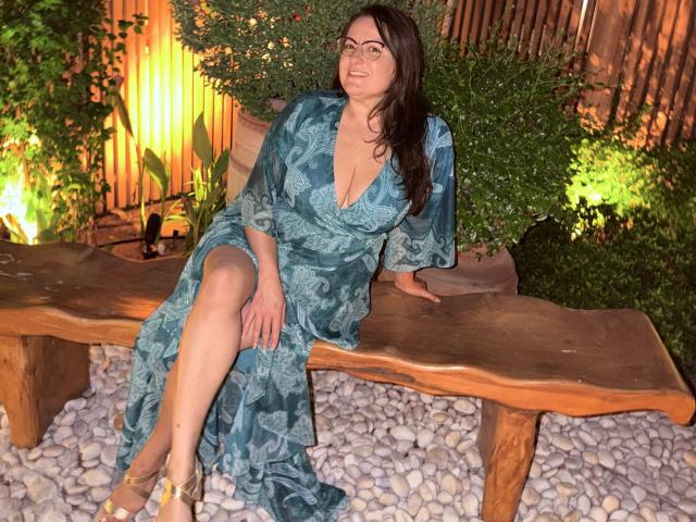BadAngel, a curvy woman with glasses and long dark hair, sits on a wooden bench, wearing a blue patterned dress, showing her tits and crossed legs, smiling.