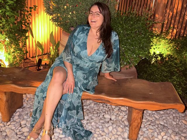 BadAngel with tits in a blue floral dress, sitting on a wooden bench, legs crossed, smiling, glasses, brown hair, pebbled ground, wooden fence, greenery, yellow sandals.