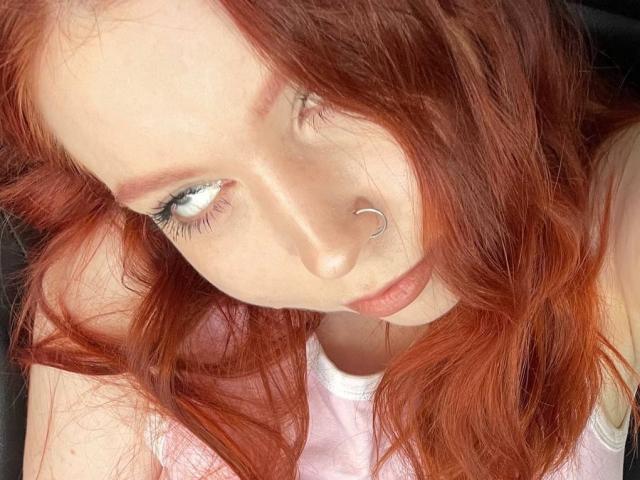 LilliamChevalier with red hair, nose ring, and blue eyes, looking up with slightly parted lips, wearing a pink top, her tits not visible, close-up selfie.
