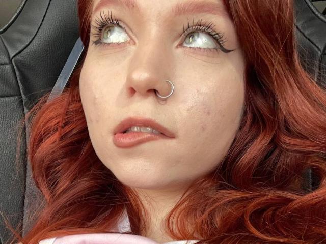 LilliamChevalier with red hair, nose ring, and big eyelashes looking up, has slightly flushed skin and a small pimple on her cheek. She's wearing pink lipstick.