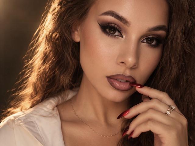 Niferttiti has sultry brown eyes, full lips with red lipstick, and long red nails. She's touching her lips with her finger, wearing a white shirt and a ring.