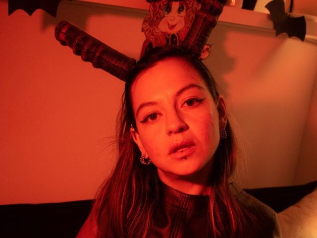 ArianaBlonde with red-tinted lighting, wearing a funny brown hat with ram horns, pouty lips, and subtle makeup, has earrings and a wavy brown-haired, cute face.