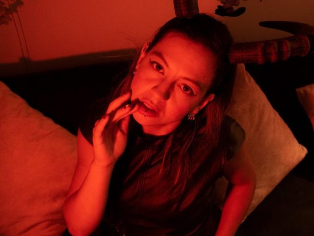 ArianaBlonde with red-lit face, wearing horn headpiece, black top, and earrings, sucking her finger, looking up, sitting on dark couch with white pillows.