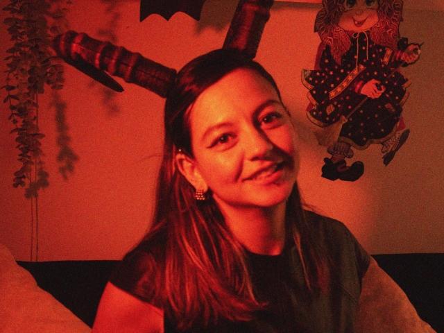 ArianaBlonde with red horns, smiling, wearing a black shirt, small earrings, sitting in dim red light, wall ornament with cute character on right.