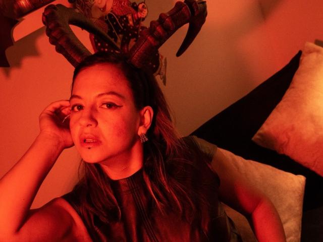 ArianaBlonde with red-lit face, wearing black horn headpiece, sleeveless top, and earrings, touching her head with a slightly open mouth, showing a small piercing on her lower lip.