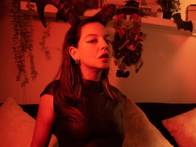 ArianaBlonde with red-tinted light, wearing a black leather top, horn headband, and earrings, sitting on a couch with pillows, looking to the side, with a doll in background.