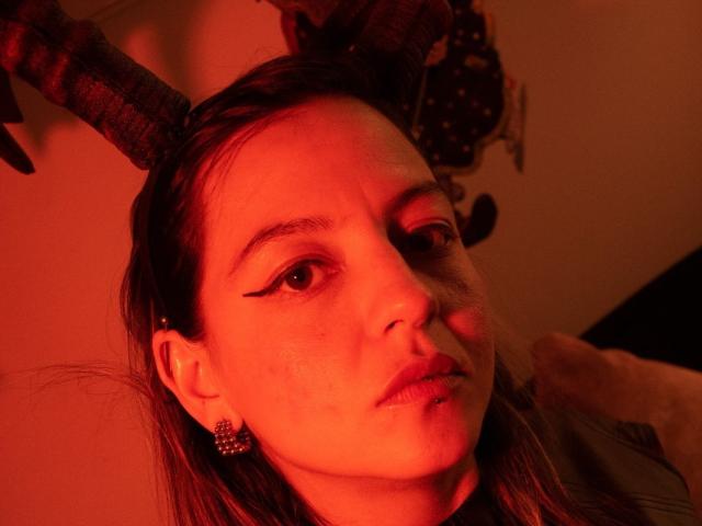 ArianaBlonde has red-lit skin, small nose, and brown eyes with winged eyeliner. She wears ram horns and pink earrings, looking slightly pouty. Her face is lit in a warm red tone.