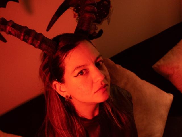 ArianaBlonde with red-horned devil headpiece, looking up with a serious expression, wearing earrings, and red lighting. Her face is slightly freckled and she has long brown hair.