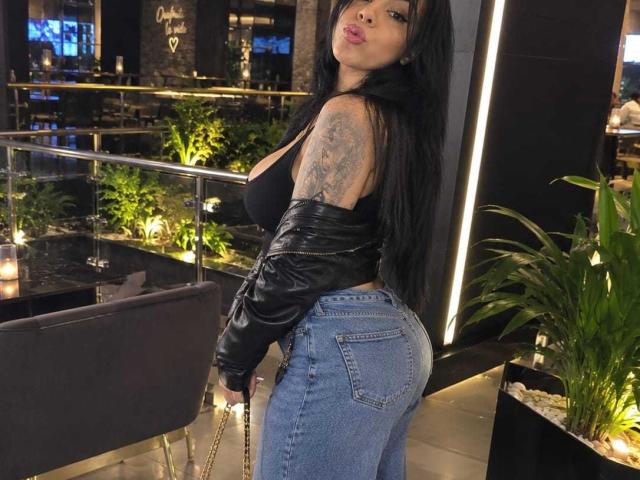 NenaBlack with long black hair, black tank top, leather jacket, and blue jeans, showing her big ass and tattooed arm, standing in a modern, dimly-lit room.