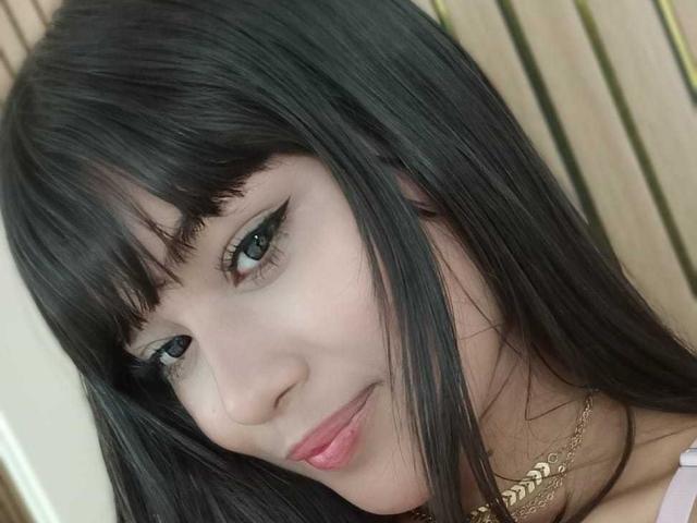 AshlyLee has long black hair, thick black eyeliner, light skin, pink lipstick, and a gold necklace, smiling subtly at the camera with green eyes.
