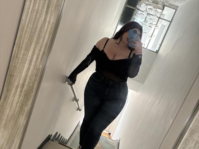 VenusxMusa con il seno prosperoso, indossa una maglietta nera a spalle scoperte e jeans neri a vita alta, sta scattando un selfie allo specchio in un corridoio stretto, tenendo in mano un telefono blu..