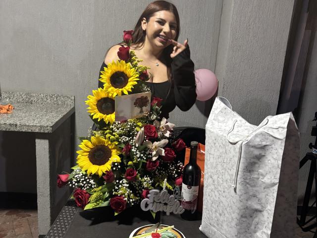 VenusxMusa mit Sonnenblumen, Rosen und roten Blumen in einem Strauß, schwarzes Oberteil, beim Essen, mit Finger im Mund, Weinflasche, weiße Tasche, Tisch mit Pizza, graue Wand, rosa Ballon..