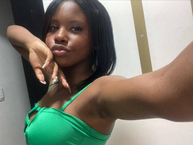 CatalinaSanA with dark skin, green tank top, and straight black hair, showing off her tits and raised arm in a selfie, looking at the camera with a slight pout.