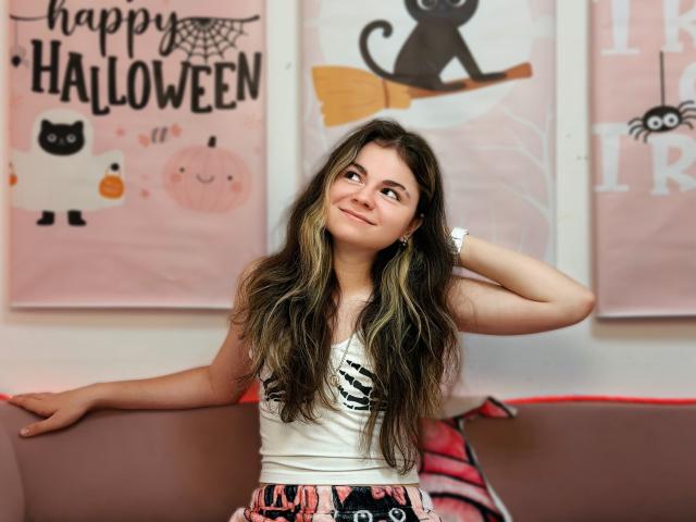 TinaRedly with long, wavy brown hair, white tank top, and pink patterned skirt, smiling, hand in hair, sitting on pink couch, wearing a silver watch.